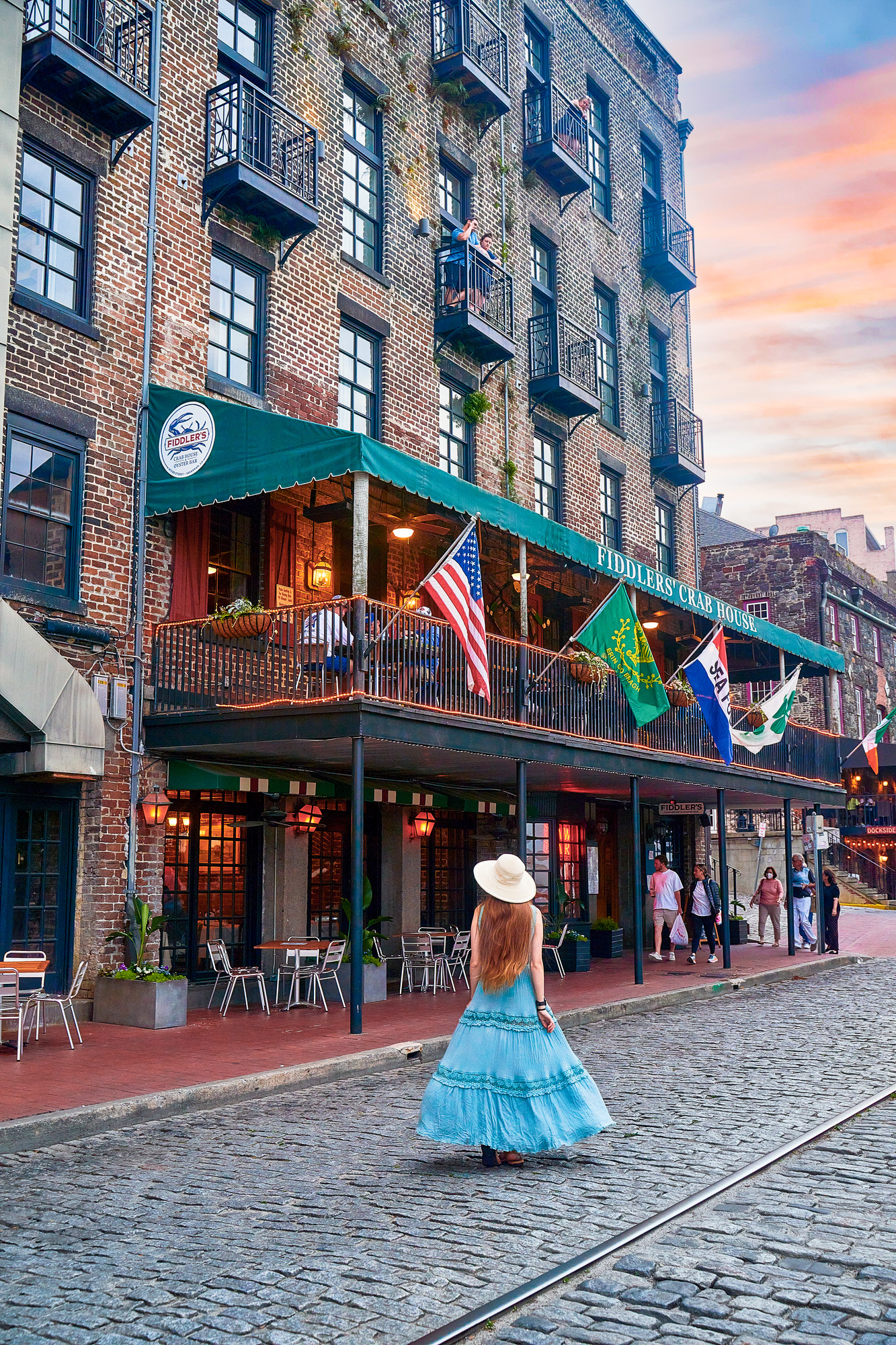 A woman in a long blue dress with her long hair down wearing a white sun hat walking on a cobblestone road. She is standing in front of a building that looks to be a pub. It is a large brick building with outdoor seating, a balcony with outdoor seating and flags, and smaller private balconies up the building. There are people standing on two of the smaller balconies. 