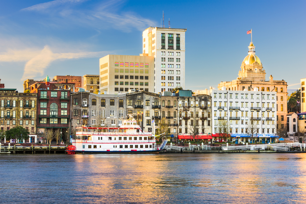 The Savannah city skyline on a sunny day. It has a mixture of old and new buildings along the river. You can also see the river and a red and white Paddlewheeler docked in front of the buildings on the river. 