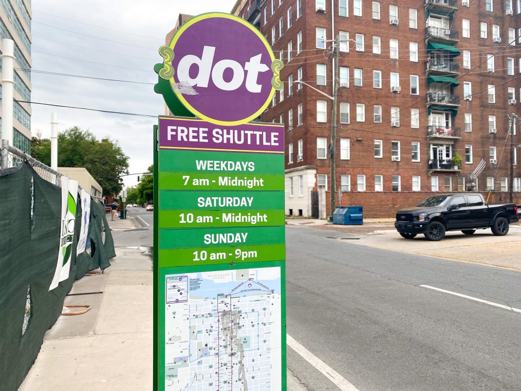 The free shuttle stop on a street in Savannah. It is a tall green sign that says "Free Shuttle" in white in a purple block. It then says the daily hours and a map of the route. On top of the green sign is a large circular attachment that is purple and says 'dot'. The street is a fairly normal street with a chain link fence by the sidewalk and a large brick apartment building across the street. 