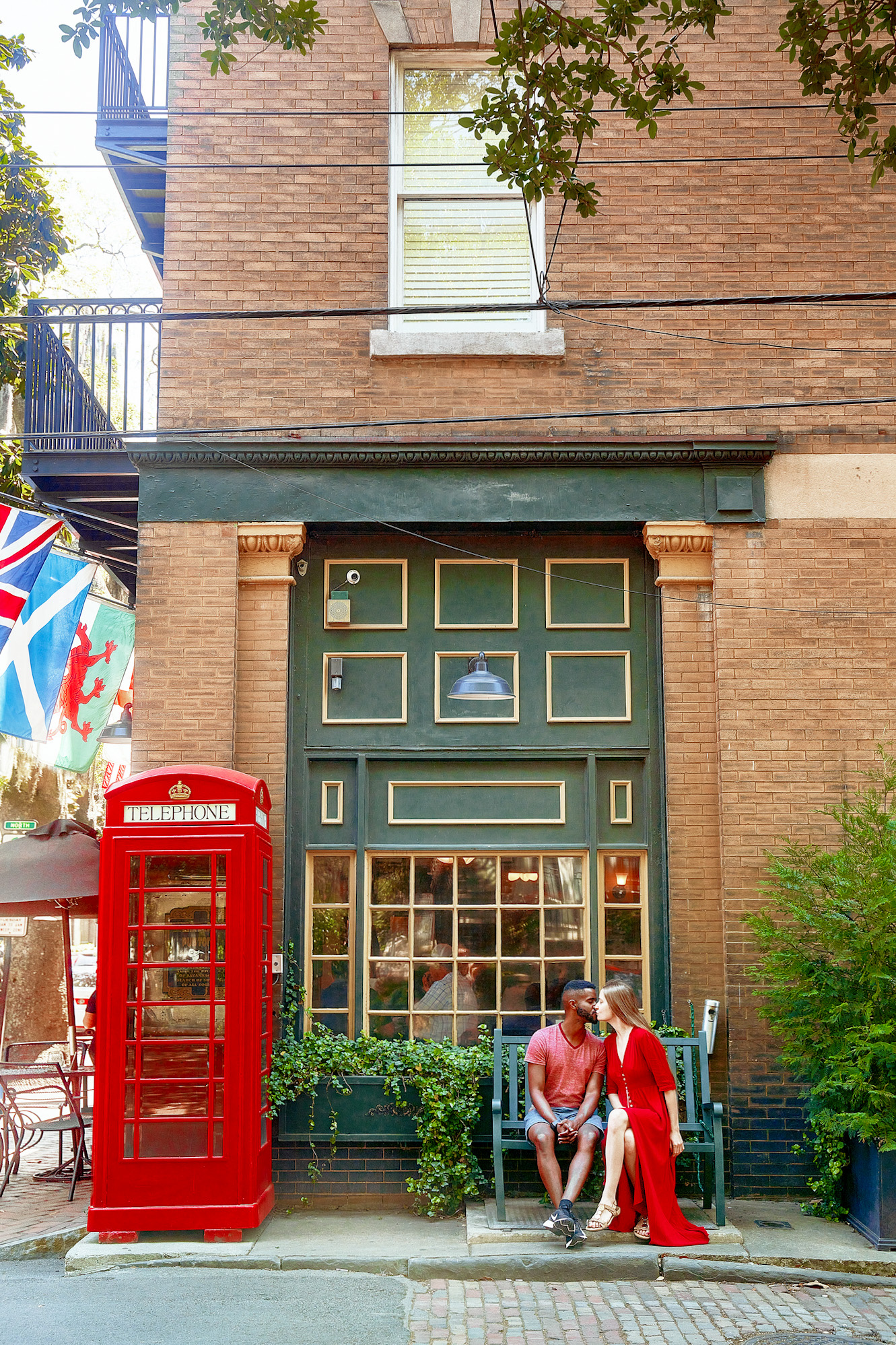 A couple sitting outside of a traditional British pub on a green bench. The Pub is mostly brick with green and yellow windows and trim. There is a classic British Telephone Booth next to a window looking into the pub. The couple is sitting on a green bench in front of the window and there is a window box with vines in front of the window. The woman in the couple is wearing a long red dress. The man in the couple is wearing a red heather shirt and denim blue shorts. They are kissing. 