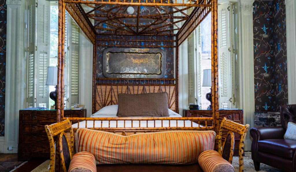 A bed with an orange bedframe that has a canopy and an orange bench in front of it with an orange and brown striped pillow. Behind the bed, is a wall with a dark wallpaper with pops of bright blue. There are two windows, each on either side of the bed. Its a great place to stay during a weekend in Charleston