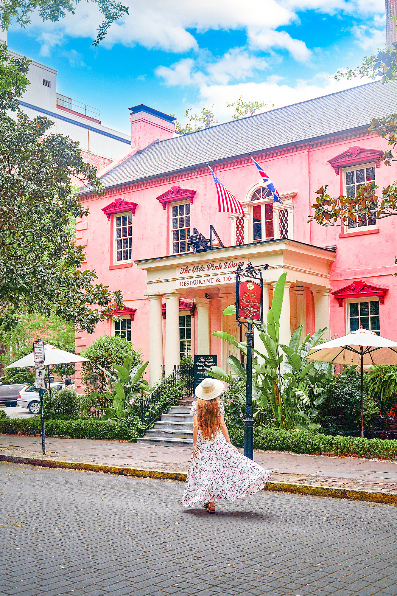 A woman standing on a cobblestone street in a long floral dress with her long hair down and a white sun hat. She is standing in front of The Olde Pink House, a historic building that is now a restaurant and tavern. It is a large building that is kind of the color of pepto bismol. The windows have a darker pink trim. There are umbrellas and lots of greenery in front of the building. A unique building to see during your 3 days in Savannah