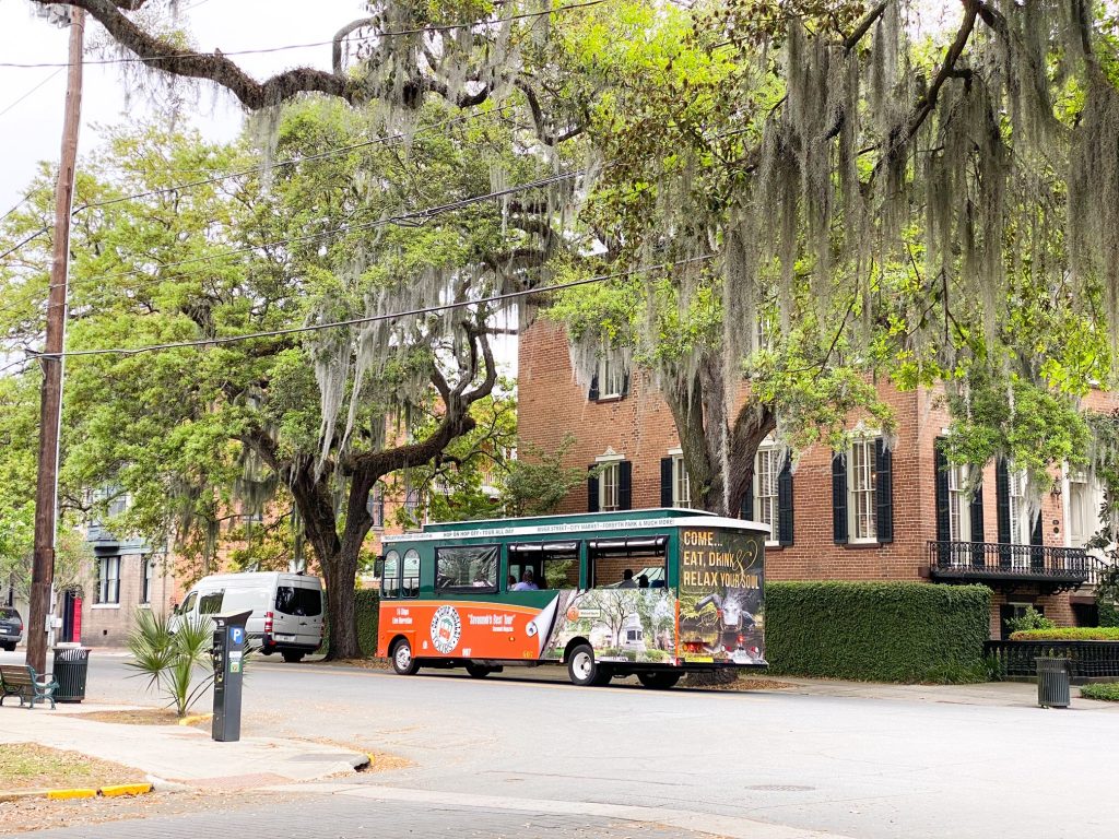 The hop-on hop-off trolley in historic Savannah Georgia. It is Green with large windows and an orange decal with images of Savannah on it. It is on a street next to a large historic brick building with lots of windows and black shutters. There are large trees covered in Spanish moss over the trolley. A fun way to get around during your 3 days in Savannah