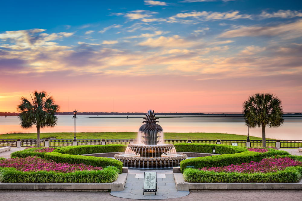 The famous Palmetto Tree Fountain surrounded by manicured shrubs and plants looking out to Charleston Harbor. There are palm tress and it is sunset.