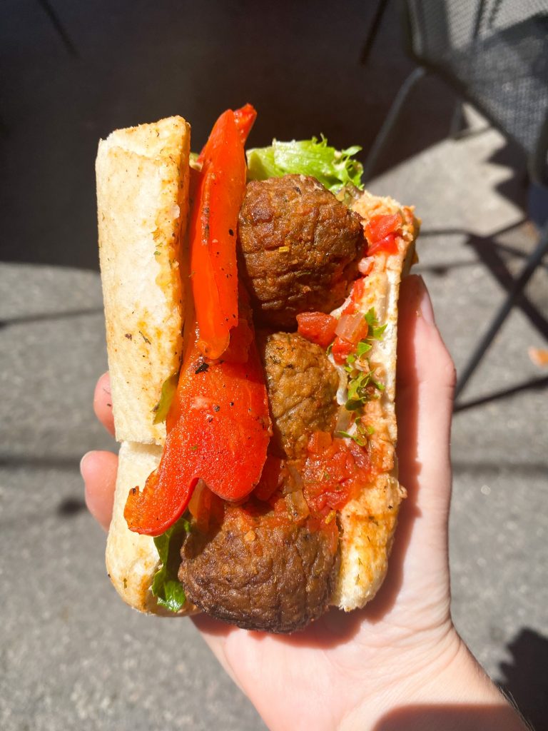 A vegetarian meatball sandwich. There are large meatballs, lettuce, red peppers, and what looks like red sauce in a Italian sub bun. Someone is holding the sandwich. 