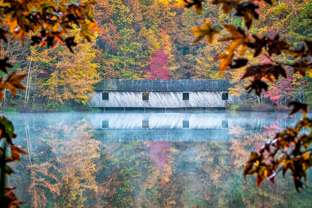 A covered bridge crosses a lake that's reflecting the Alabama fall foliage.