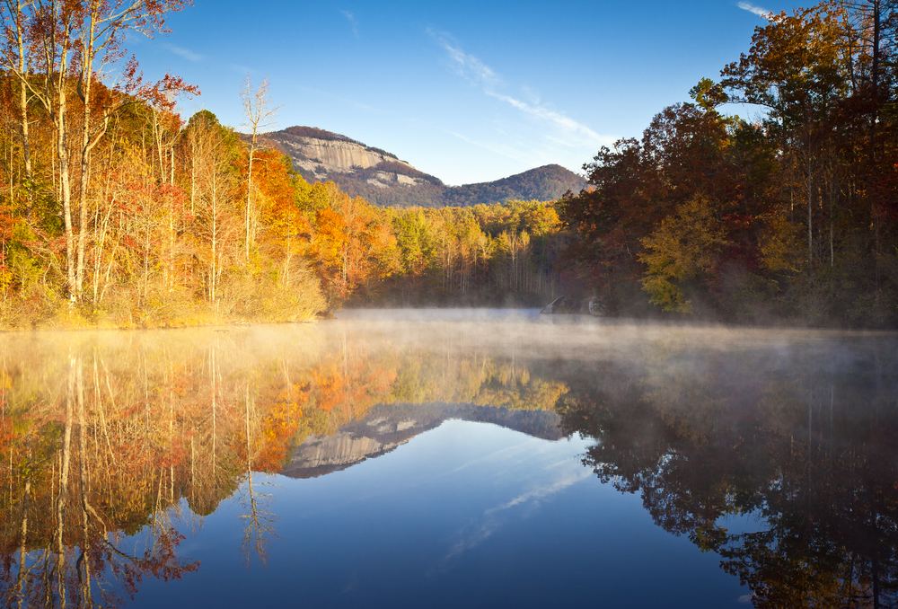fog-covered lake with a reflection of fall colored trees and Table Rock Mountain in the background.