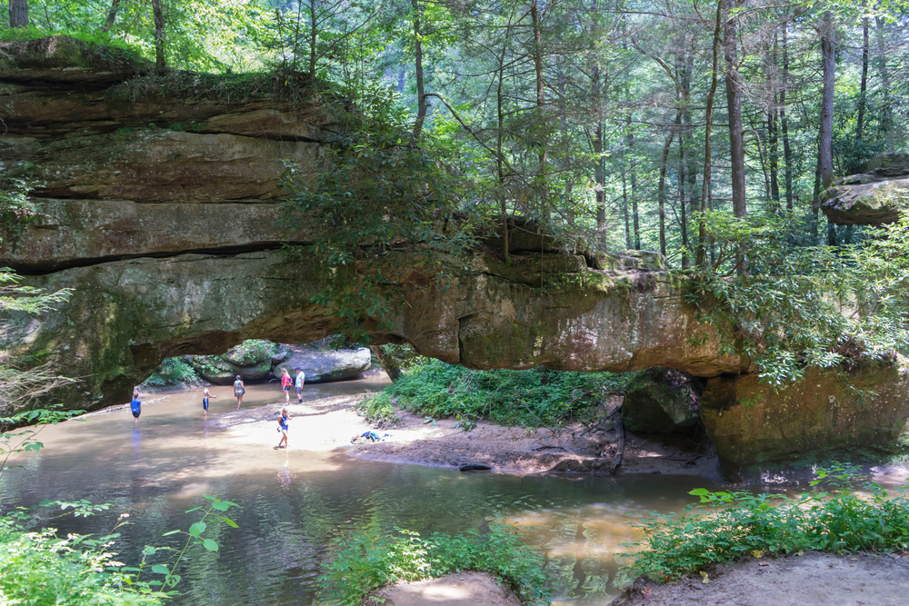 Photo of Rock Bridge spanning across a small river with people walking on the river bank below in the distance.