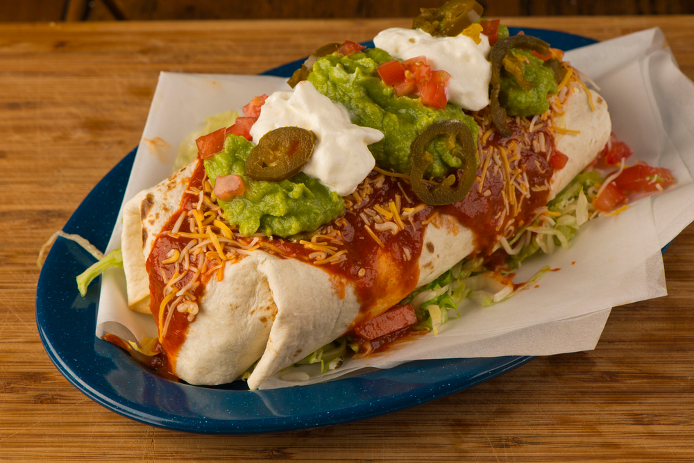 A tex Mex burrito served on blue plate topped with cheese, salsa, guacamole sour cream and jalapeños