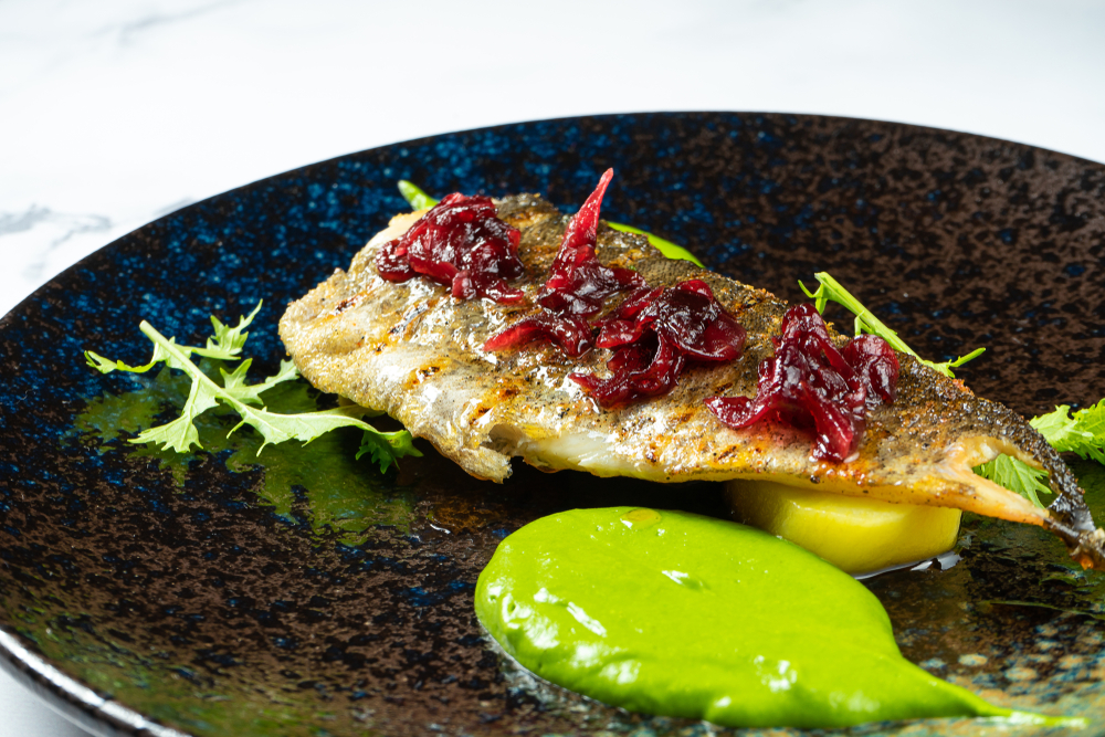 A beautiful fish dish with a green sauce and veggies on a sparkly plate