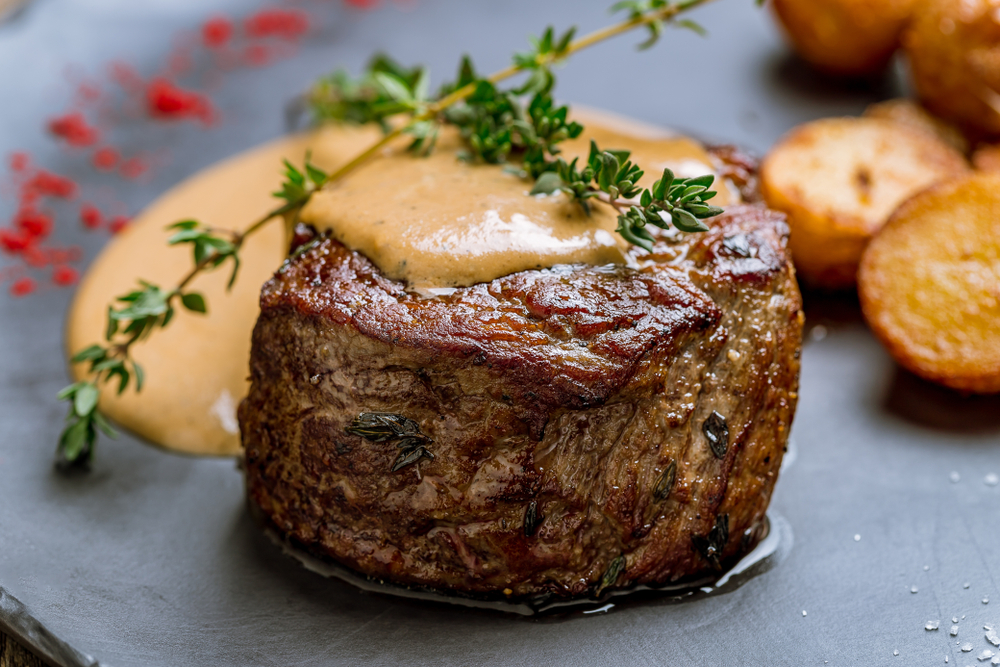 A filet steak topped with sauce and twig of rosemary and potatoes in background on a black plate