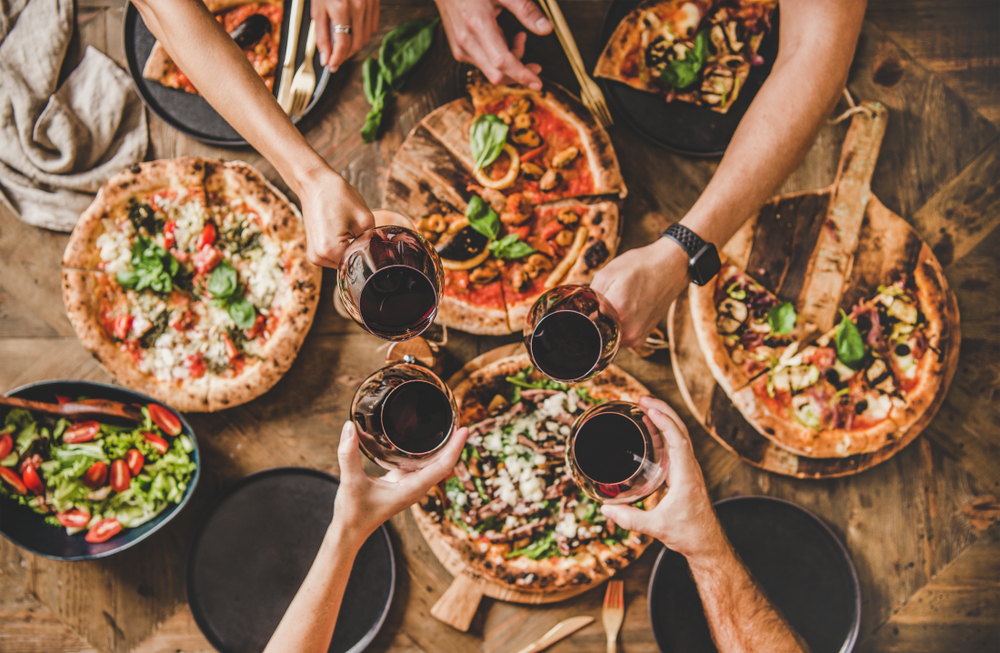 A photo of extended arms holding full red wine glasses over several delicious pizzas.