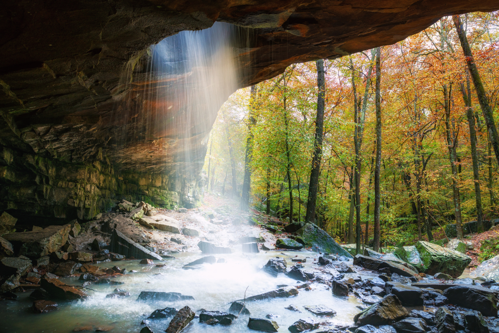 A photo of a Glory Hole Falls, a waterfall in Arkansas, falling through a hole created in a bluff into a rocky pool of water with autumn trees behind it.
