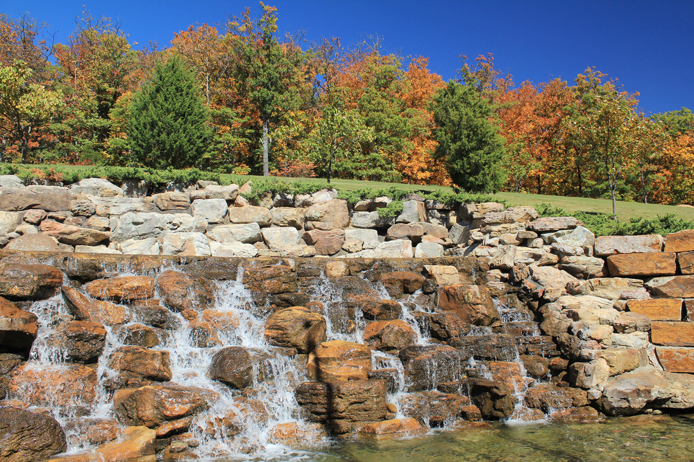 A picture of a rocky waterfall in front of a forest of trees with fall colored leaves on a sunny day.