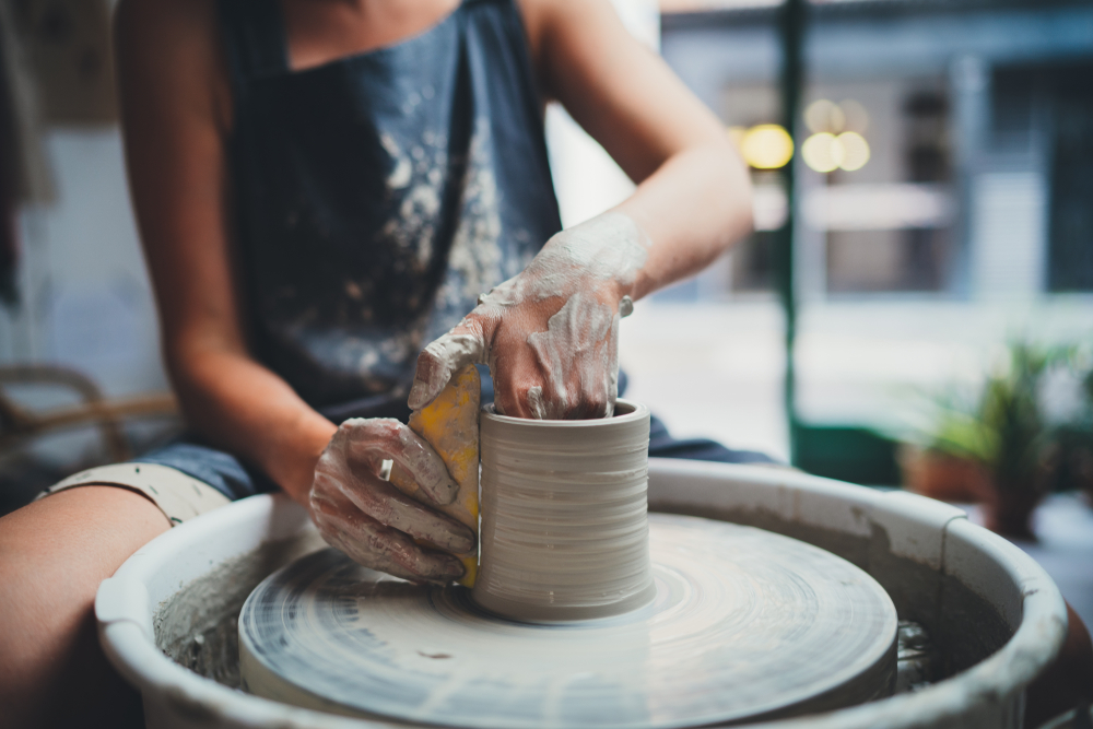A person making a cup or vase on the pottery wheel with white clay. They have one hand in the cup and the other outside it holding a flat item up to the cup. Their hands are covered in wet clay. They are wearing a blue apron that is also covered in wet clay. 