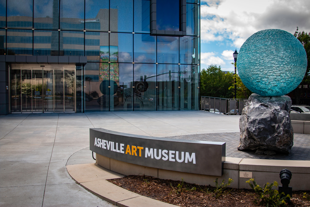 The front of the Asheville Art Museum, a great spot during a weekend in Asheville. It is a large building that looks made of glass windows. In front there is a patio area with a sign for the museum and a sculpture with a large boulder and a blue glass orb balanced on top of it. 