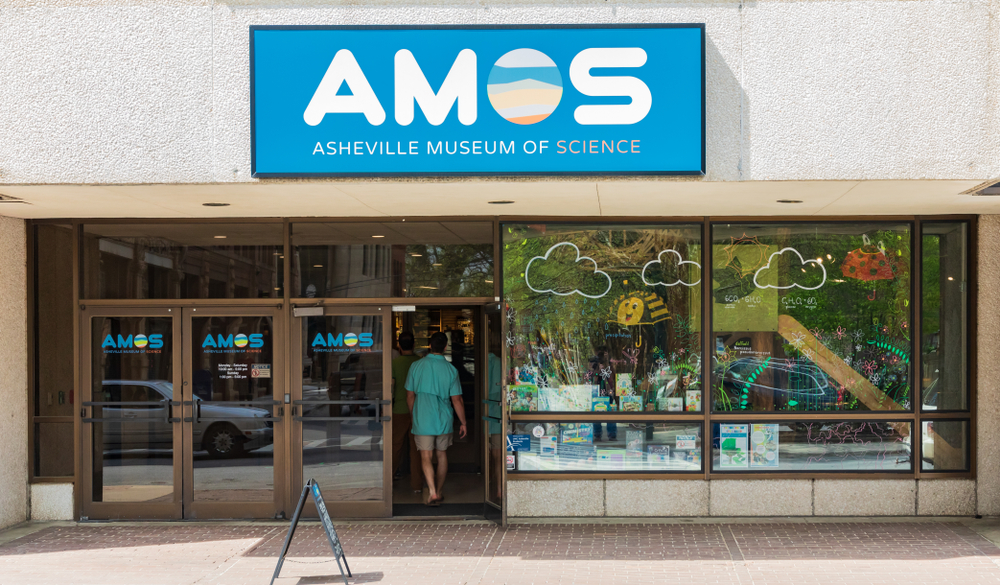 The exterior of a concrete building with two sets of double doors and some windows in front of it. Over the doors and windows is a sign for the Asheville Museum of Science. There is a person walking through the door and displays in the windows. 