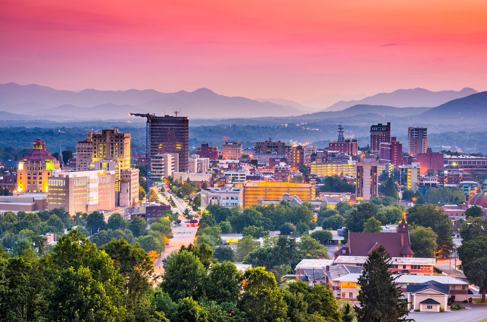 A view of the Asheville skyline. It is twilight so the sky is pink, purple and blue. At the front of the image you can see the skyline with buildings all lit up and trees scattered around the city. Behind the city you can see the Blue Ridge Mountains which are almost in silhouette in a pale dark blue and purple. 