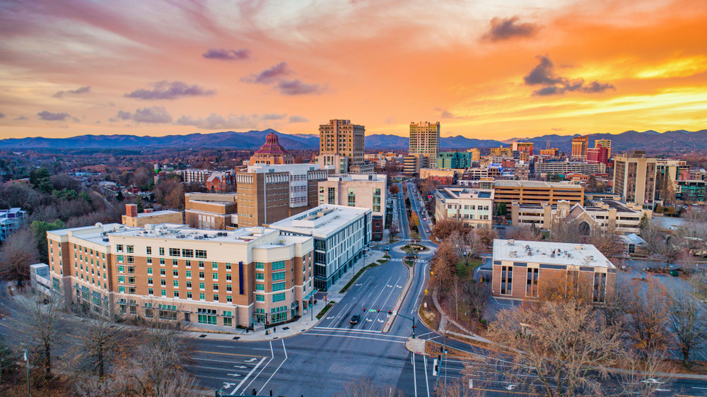 an aerial view of Asheville at sunset. The sky is orange and yellow. You can see buildings, streets, and the mountains in the distance on your weekend in Asheville.