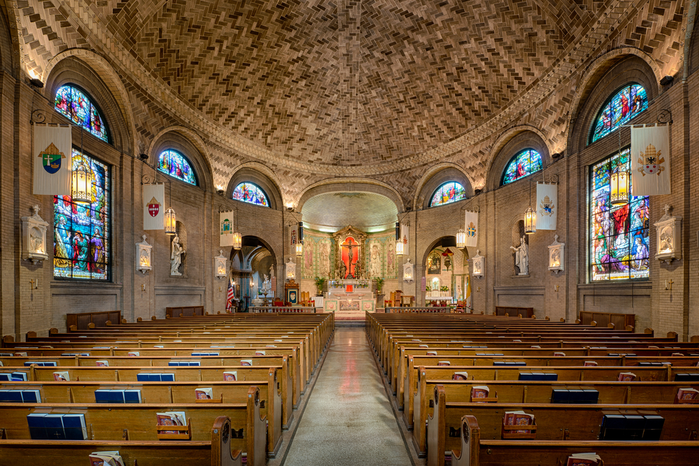 The inside of the Basilica of Saint Lawrence. The ceiling is herringbone laid brick, there are large stained glass windows on either side of the building, and at the end there is an elaborate alter. There are rows of wooden pews with bibles and hymnals in them. 