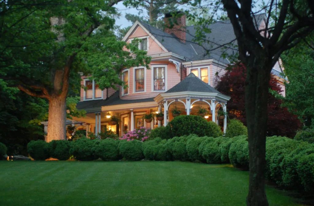 A historic Victorian style home. It is painted a peachy pink color and has white trim. In front of it there is a green lawn, large trees, and shrubs. Some of the shrubs have pink flowers. On the porch you can see ferns hanging from the ceiling and lights on. It is one of the best places to stay during a weekend in Asheville. 