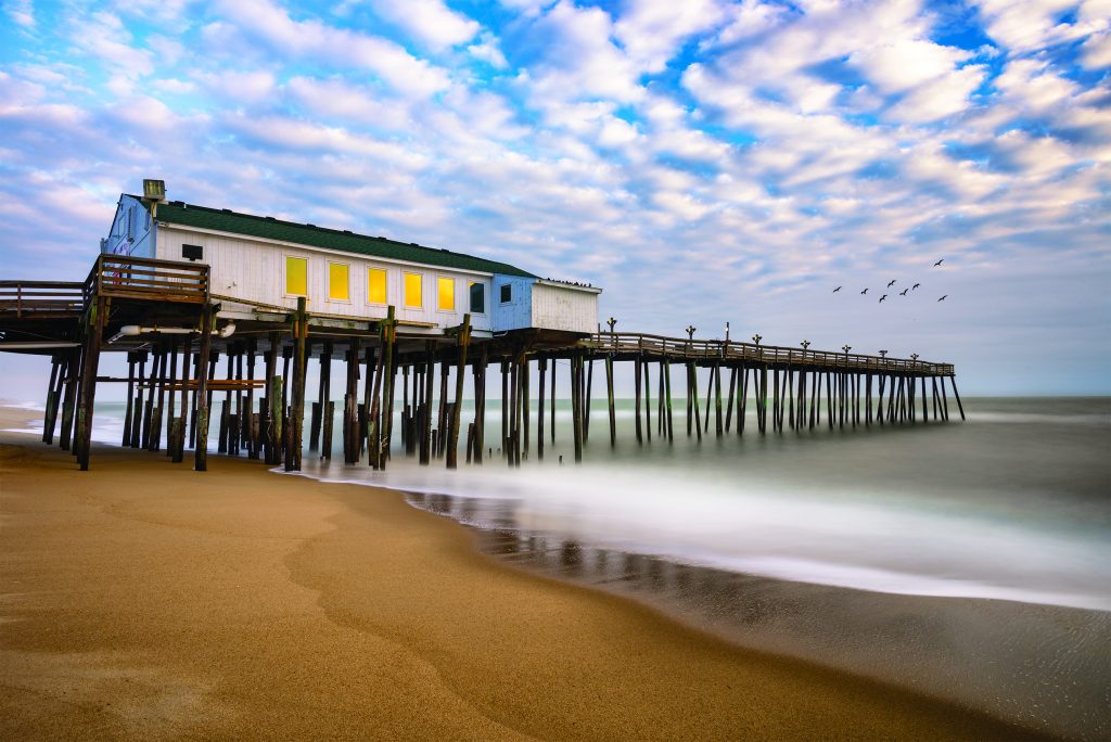 The pier at Kitty Hawk on the Outer Banks stand over misty waves washing up on shore.