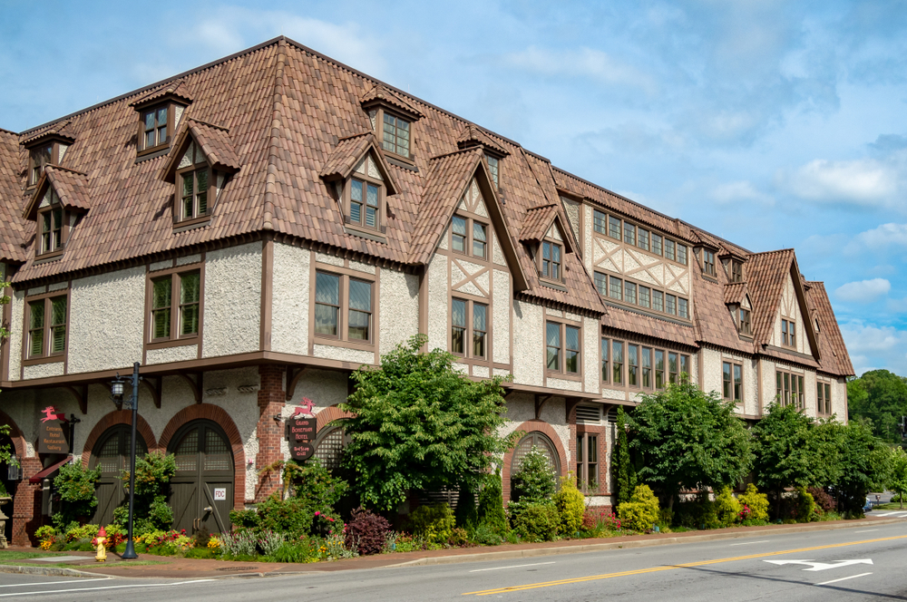 The exterior of a set of buildings at the Biltmore Village. The buildings are styled like old European cottages with the tile on the roof supposed to look like a thatched roof. It is a cream color with brown trim around the doors and windows. In front of it there is a garden that wraps around its exterior where there are shrubs and plants with red flowers. 