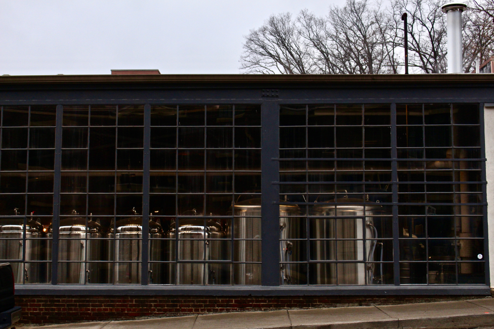 The exterior of a dark blue building that has large windows in front of it. Through the windows you can see silver tanks that are used for brewing beer. 