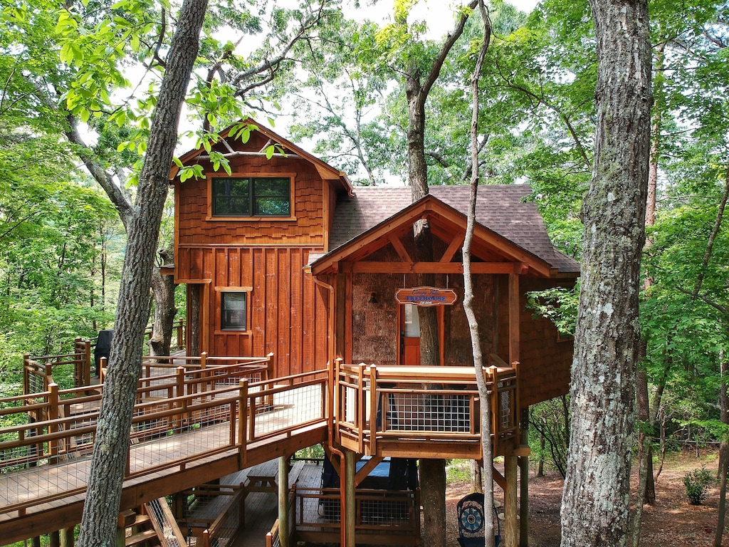 A wooden treehouse on a platform in the woods. It has a ramp leading to a large porch that wraps around the treehouse. You can see trees coming through the deck and the house. 