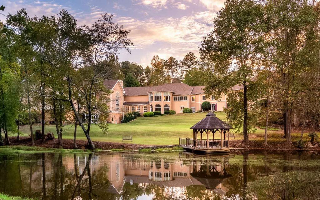 The view of the Château Élan Winery & Resort. It is a large classic style building that is kind of peach in color. In front of it there is a large pond with green algae, trees around it, and a gazebo on the shore.