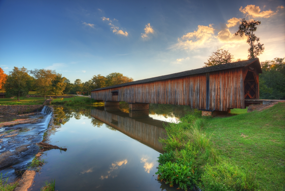 A long wooden covered bridge. It connects two pieces of grassy land across a river. The river has a small manmade waterfall not far from the covered bridge. 