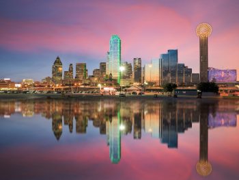 The Dallas skyline at twilight from the view of the river. The skyline is all lit up and the sky is mostly pink and purple with some blue. The sky and the skyline is reflected in the river. Best day trips from Dallas.