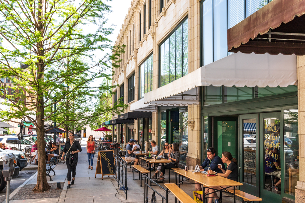 Looking down the sidewalk on a street in Downtown Asheville. There are people sitting at tables outside of various restaurants that line the street. The restaurants all have awnings over them and their own section of seating. There are also people walking down the sidewalk. 