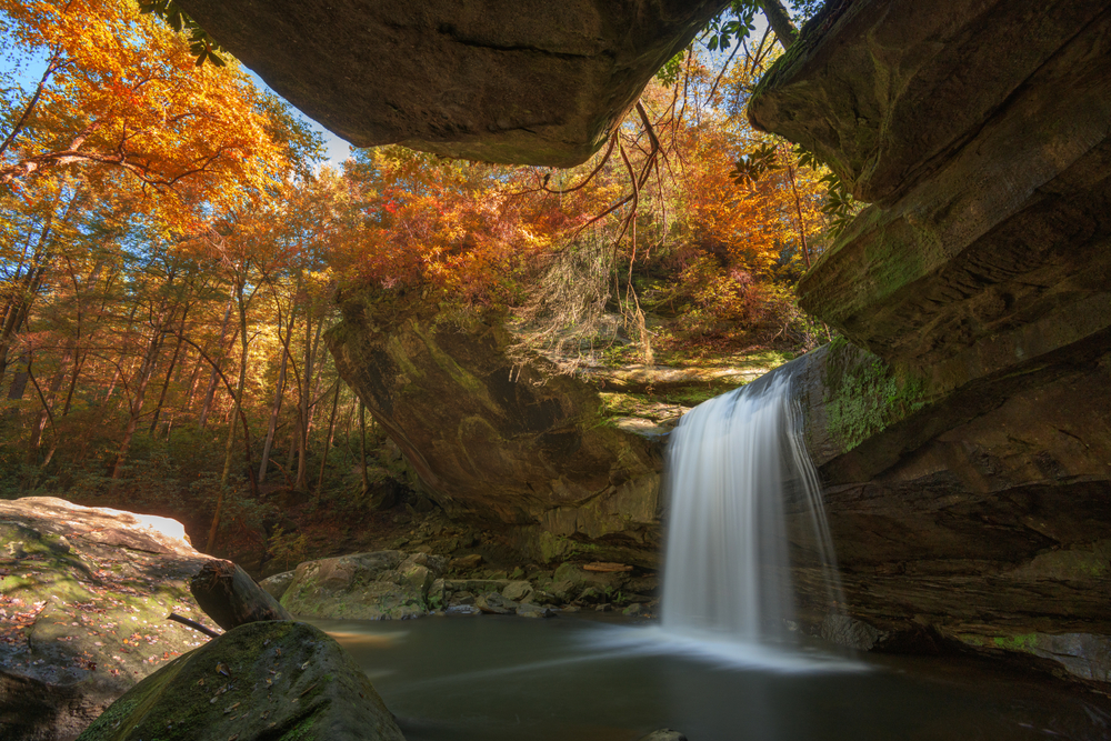 view of the waterfall from underneath a rock ledge, fall colored leaves and trees surround the rocks around the waterfall