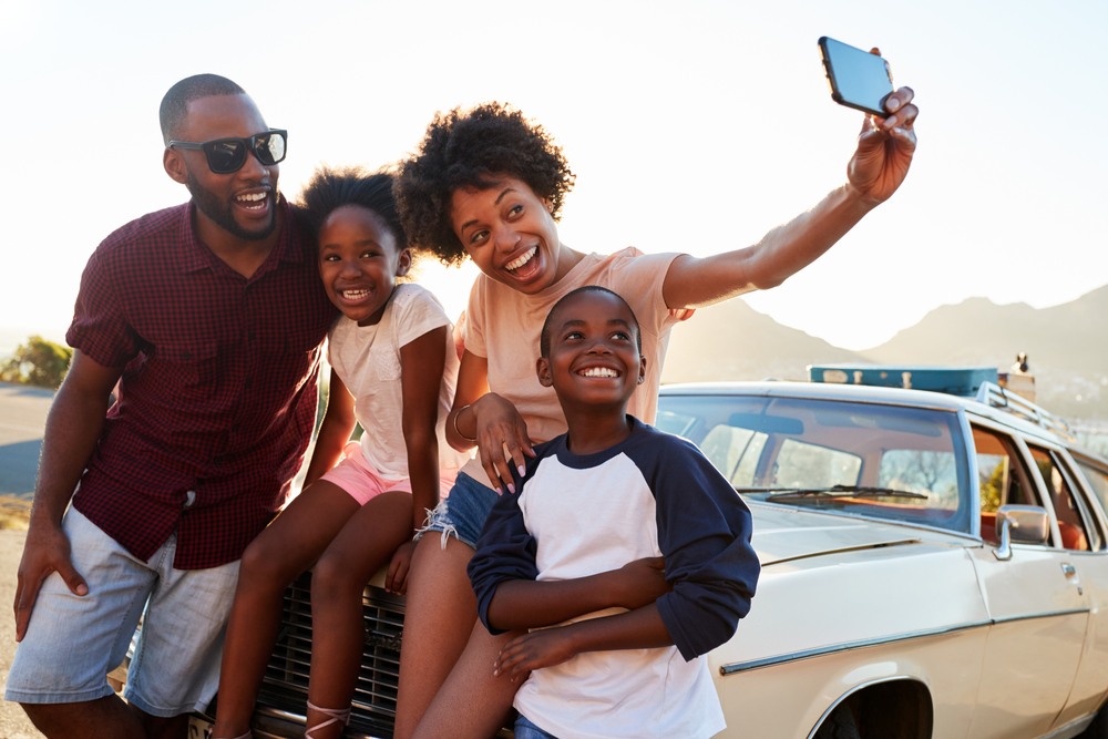 A family consisting of a man, woman, and a son and daughter. They are sitting on the hood of their car and the mom is taking a selfie. You can see luggage strapped to the top of their car.