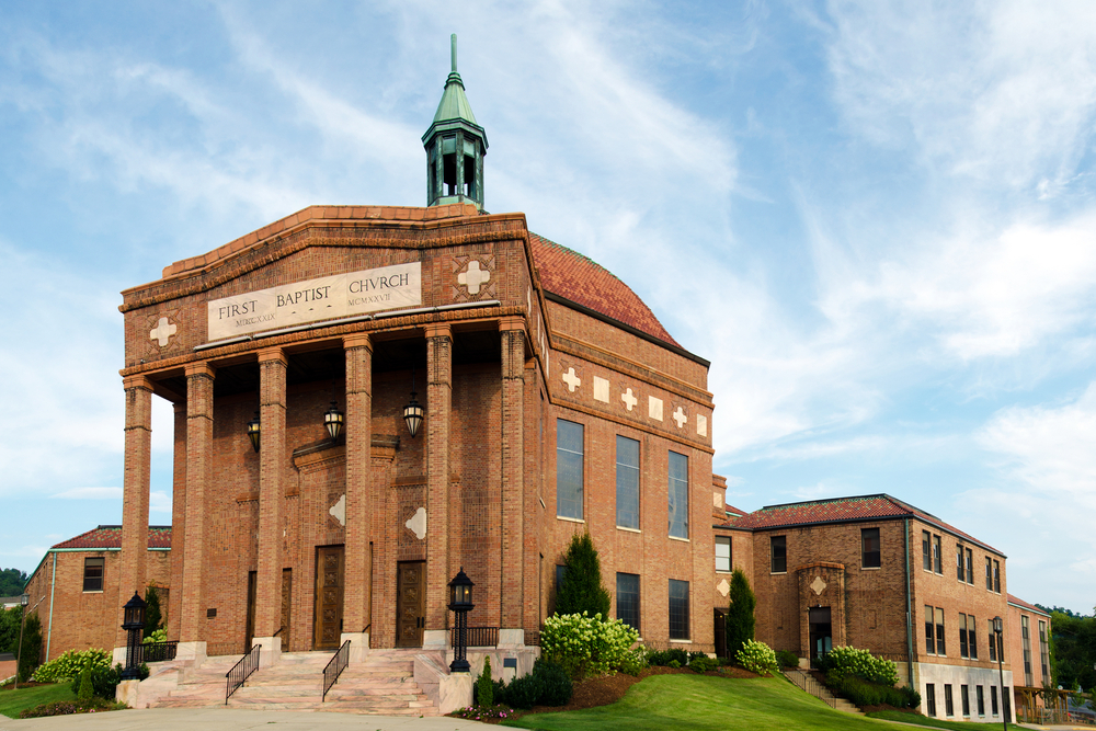 The exterior of the First Baptist Church. It is a large brick building with a brick dome in the back and a tall brick entry way on the front. There is a stone sign in front at the top of the building that says 'First Baptist Church'. 