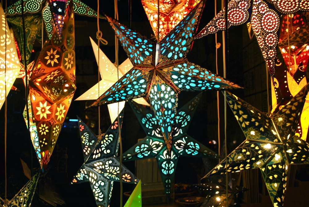 Hanging paper star lanterns in all types of colors and patterns. They are hanging from the ceiling in a dark room and are lit up from inside. 