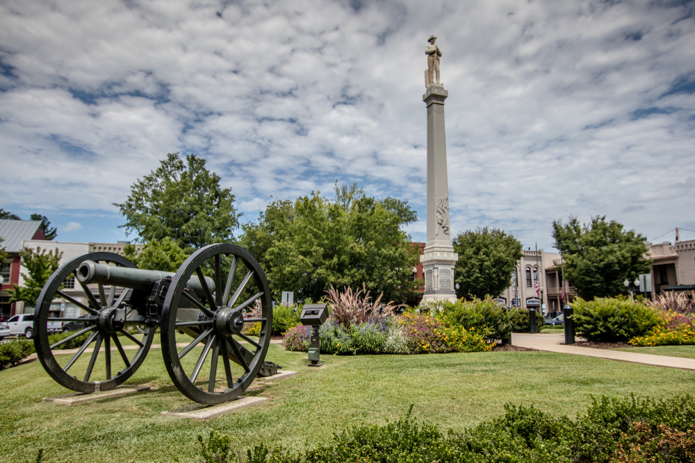 A Civil War monument in the middle of the town square of Franklin. There is a grassy area around the monument and flowers and shrubs with yellow, pink, and purple flowers. There is also an old cannon in the grassy area.