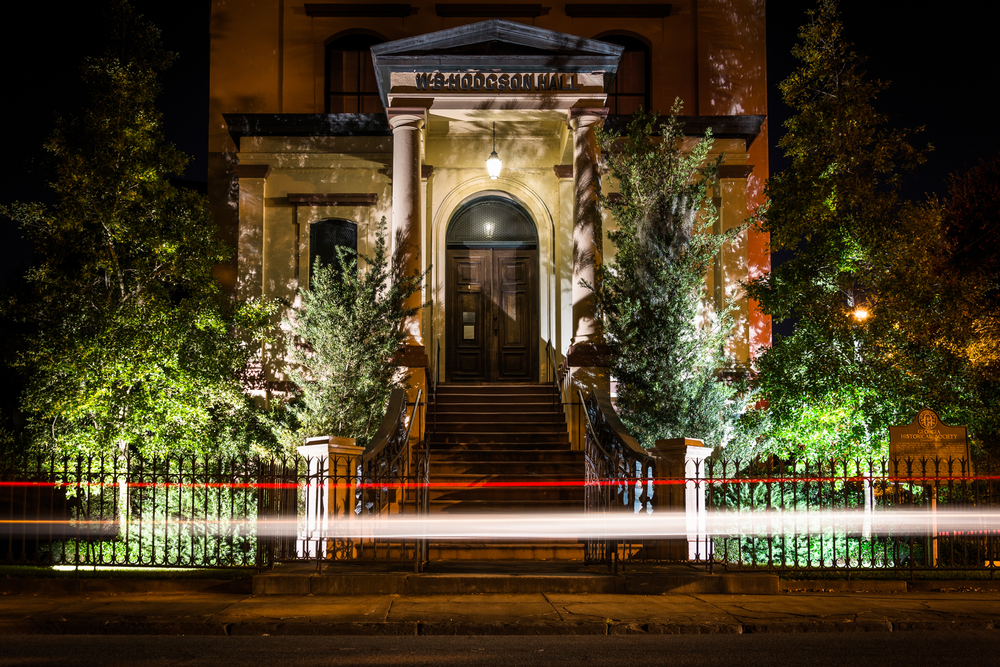 The front of a historic building in Savannah at night. The front of the building is lit up and there are two light trails, one red and one white, streaking in front of the building across the whole picture. Visiting here is one of the best things to do in Georgia 