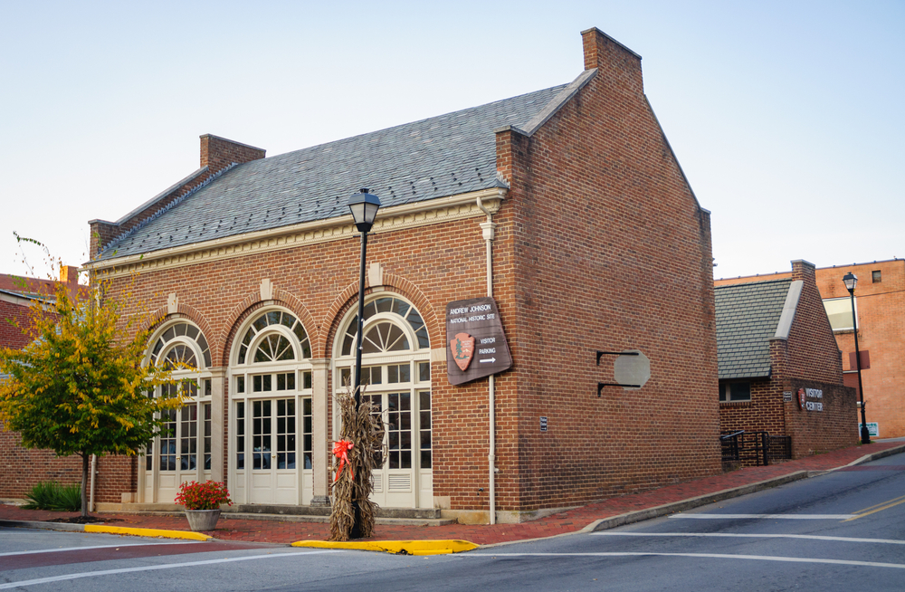 The exterior of the Andrew Johnson Historic Site. It is a brick building with tall windows on the front that arch. It is on the corner of a street and there is a smaller brick building behind it. 