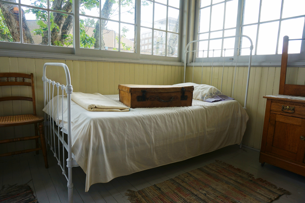 A room that looks old. There is an antique iron bed with cream sheets and an old leather suitcase is on top of it. In the room you can also see a chair, a dresser, and a rug. The walls are painted pale yellow half way up and then there are windows.