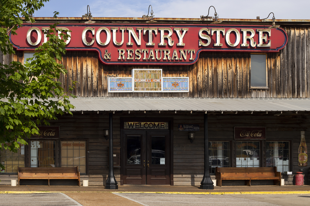 The front exterior of an old wood building with a red sign that says 'Old Country Store and Restaurant'. There are benches under an awning next to the front door. 