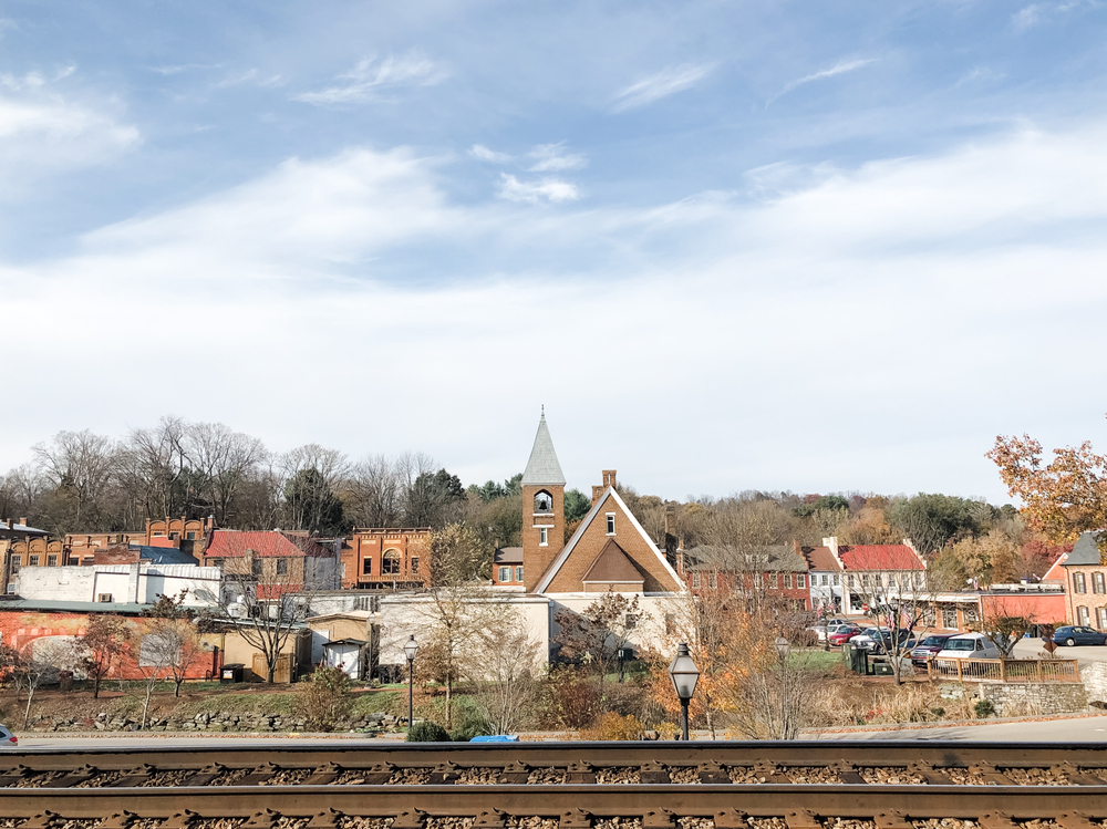 The view of a small town from across the train tracks. The buildings are smaller and made of brick and stone. They are all connected in a row. There is also a brick church with a steeple. 