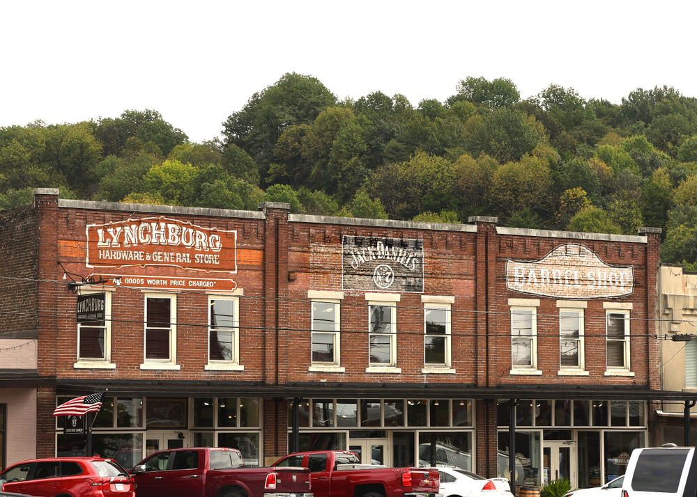 A row of old brick buildings with painted signs on them. They say 'Lynchburg Hardware and General Store', 'Jack Daniels', and 'Barrel Shop'. There are tall windows on the second floor and shop windows on the bottom of the buildings. There are red and white cars parked in front of it. 