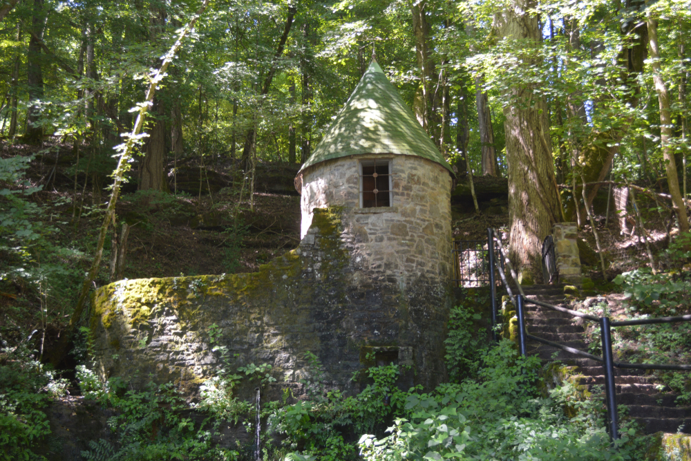 An old stone turret and wall surrounded by a dense forest. There are stairs leading up a hill with a black metal railing next to them. The stone building and wall has ivy and moss growing on it. 