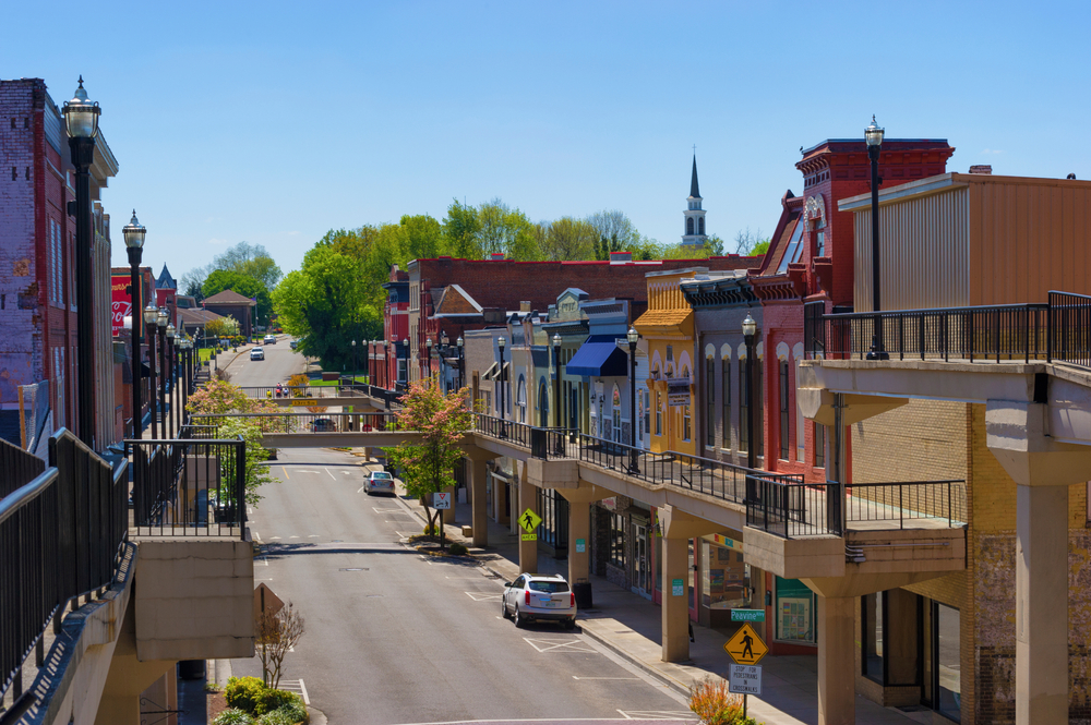 Looking down the road of a main street in the town Morristown. The fronts of the buildings are all painted different colors. There are yellow, black, blue, red, and green building fronts. They are all connected and there are trees with green leaves towards the end of the road. 