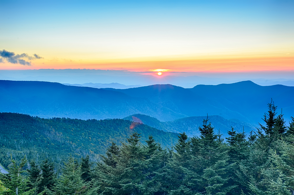 The view looking out at the Blue Ridge Mountains from the peak of Mount Mitchell. You can see mountain peaks in the distance and the sun is setting behind them. The mountains seem to be blue and are covered in trees. 