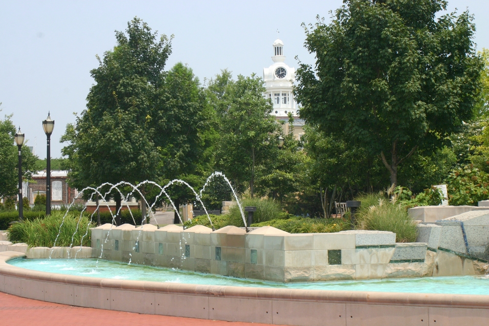 A fountain in the town square of Murfreesboro. The fountain is stone and there is water shooting out if it. Around the square there are shrubs, grasses, and green trees. There are also some light posts and you can see a government building behind the trees. 