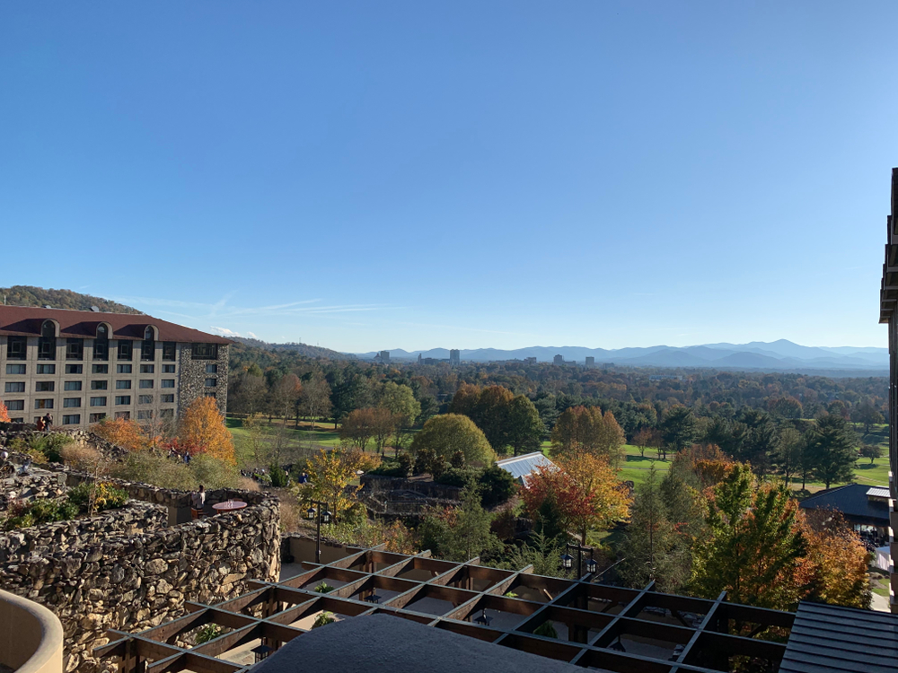 The view looking out at the Omni Grove Park Inn landscape. You can see a bar area directly below, small buildings, a green space, lots of trees, and a corner of a large stone hotel. In the distance you can see more trees and the mountains. The trees have green, red, orange, and yellow leaves. 