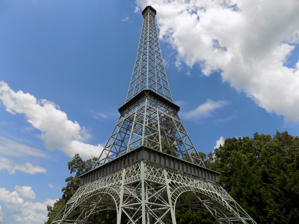 Looking up at a replica Eiffel Tower. It is a pale sage green. There are trees with green leaves behind part of it. The sky is blue and there are fluffy clouds. 